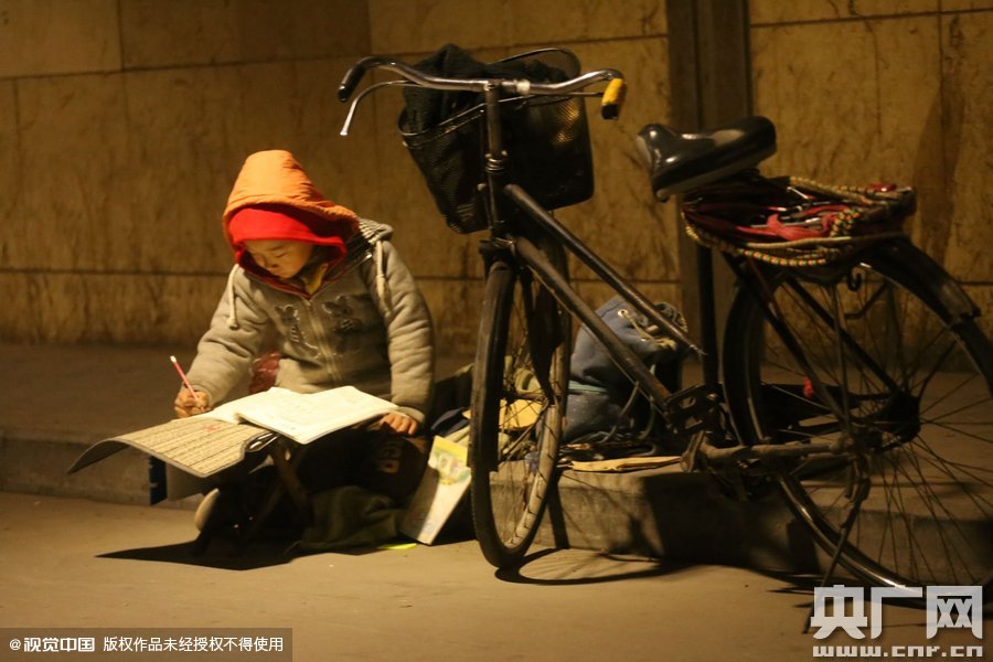 鄭州中原路京廣鐵路大橋下涵洞的馬路牙子邊，一位戴著紅色辣椒帽的小男孩趴在一個(gè)小馬扎上，上面鋪著一張車墊，在微弱的燈光下寫作業(yè)