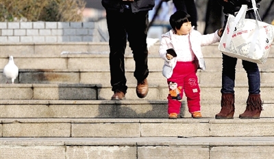 昨天中午，一女孩拉著家長的手在紫荊山公園曬太陽，氣溫升高，孩子的棉衣解開了扣子。